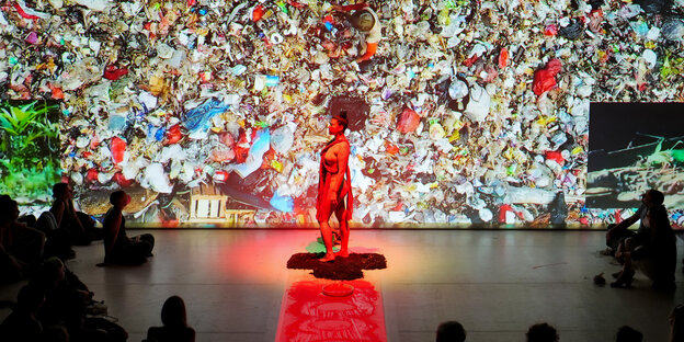 Bühnenperformance mit einer roten angeleuchteten weiblichen Figur, das Bühnenbild zeigt einen Berg Plastikmüll.
Bilduntertitel: "In Martha Hincapié Charrys Performance „Amazonia“ geht es um das Umweltbewusstsein Indigener. Foto: Gianni Bettucci"