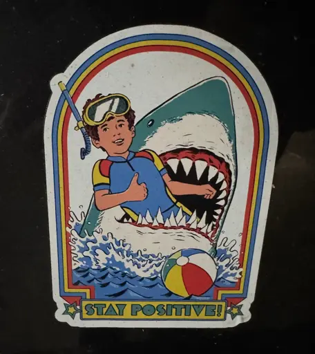 A colorful 80s cartoon inspired sticker of a child in swim gear getting eaten by a shark. The child is giving a thumbs up, with the text ‘STAY POSITIVE!’ beneath them  